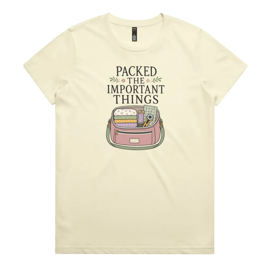 A pale butter yellow short-sleeve crewneck t-shirt featuring a graphic print on the front with the text 'PACKED THE IMPORTANT THINGS' above an illustration of a pink bag filled with quilting supplies.