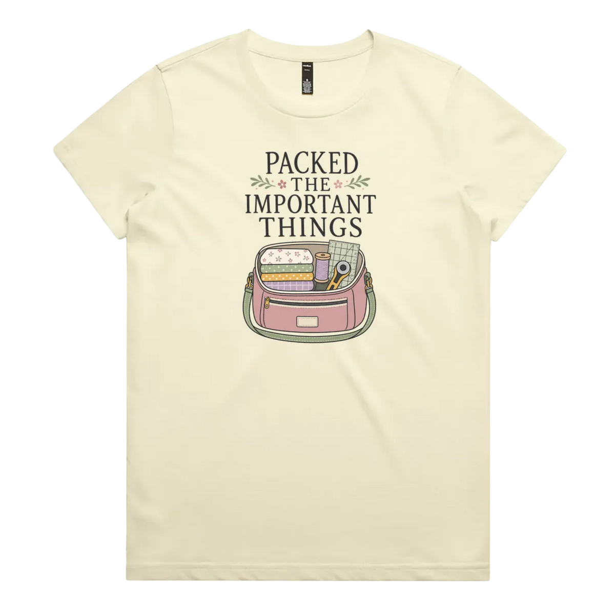 A pale butter yellow short-sleeve crewneck t-shirt featuring a graphic print on the front with the text 'PACKED THE IMPORTANT THINGS' above an illustration of a pink bag filled with quilting supplies.
