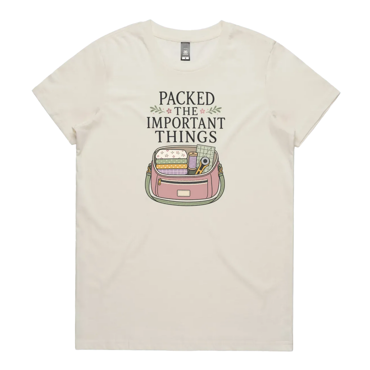 An off-white short-sleeve crewneck t-shirt featuring a graphic print on the front with the text 'PACKED THE IMPORTANT THINGS' above an illustration of a pink bag filled with quilting supplies.