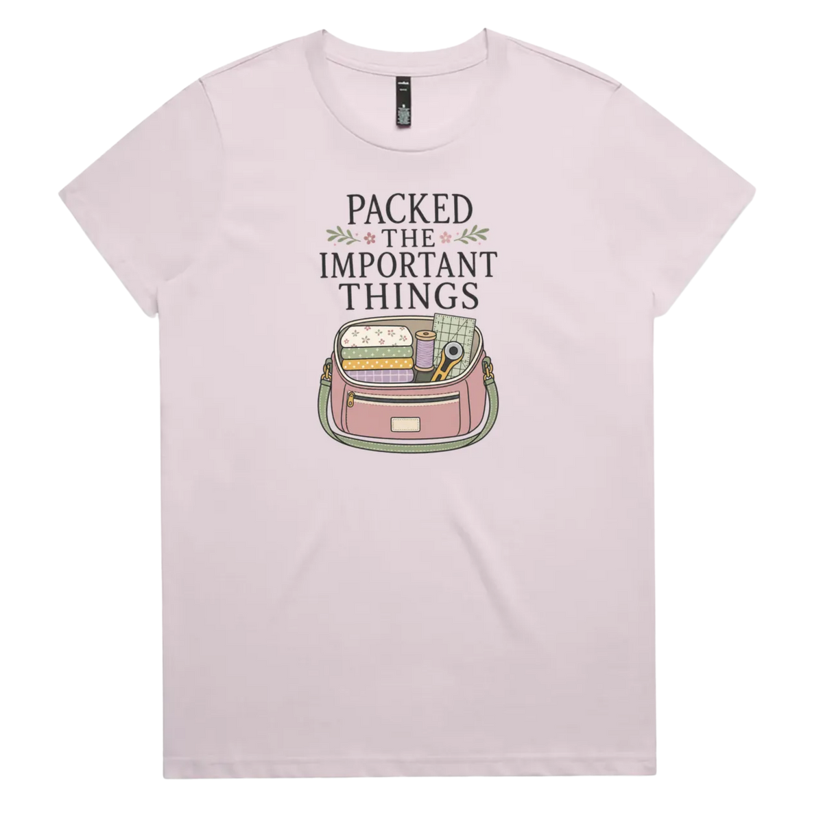 A pale mauve short-sleeve crewneck t-shirt featuring a graphic print on the front with the text 'PACKED THE IMPORTANT THINGS' above an illustration of a pink bag filled with quilting supplies.