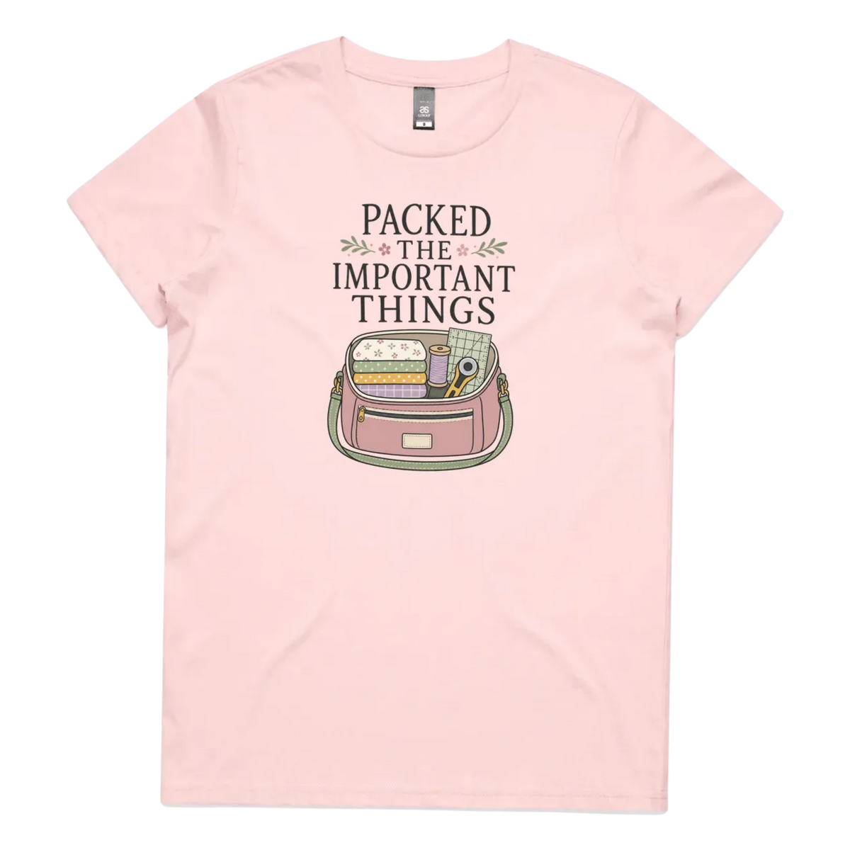 A pale pink short-sleeve crewneck t-shirt featuring a graphic print on the front with the text 'PACKED THE IMPORTANT THINGS' above an illustration of a pink bag filled with quilting supplies.