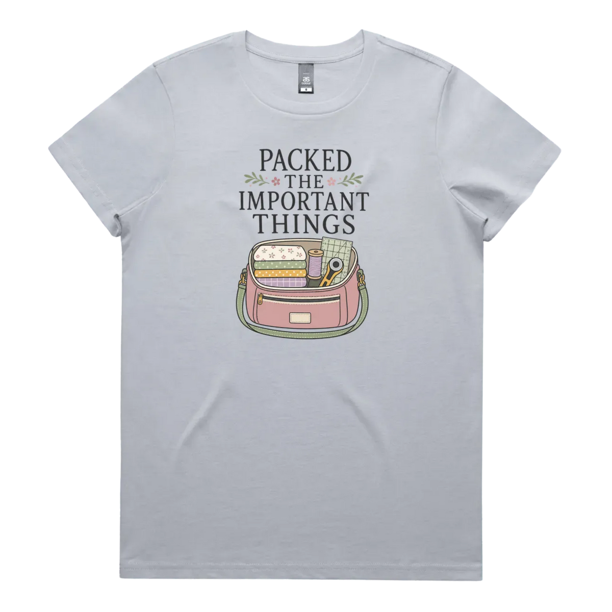 A powder blue short-sleeve crewneck t-shirt featuring a graphic print on the front with the text 'PACKED THE IMPORTANT THINGS' above an illustration of a pink bag filled with quilting supplies.