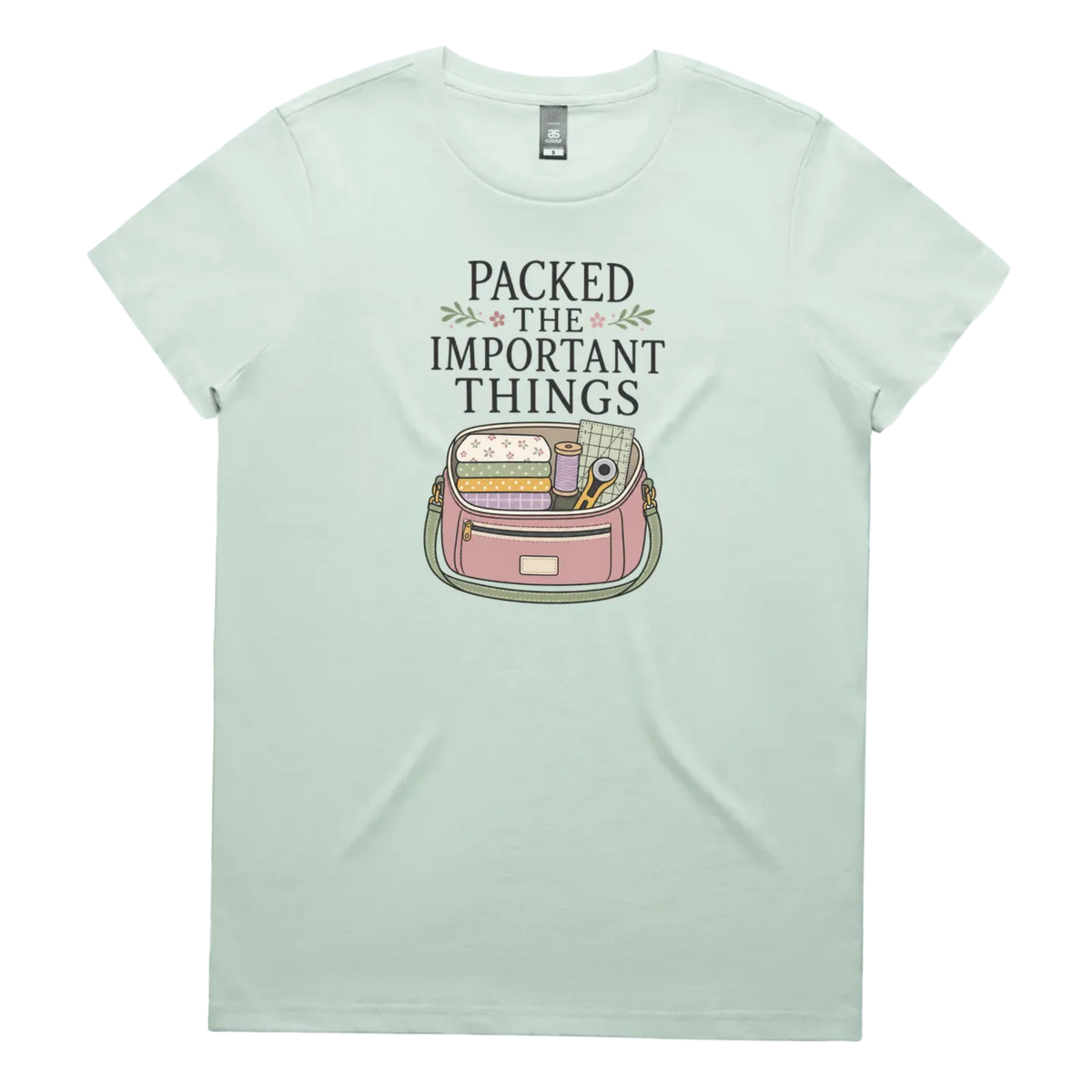 A pale mint green short-sleeve crewneck t-shirt featuring a graphic print on the front with the text 'PACKED THE IMPORTANT THINGS' above an illustration of a pink bag filled with quilting supplies.