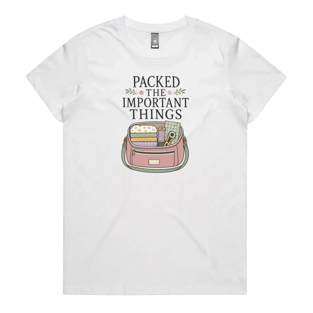 A white short-sleeve crewneck t-shirt featuring a graphic print on the front with the text 'PACKED THE IMPORTANT THINGS' above an illustration of a pink bag filled with quilting supplies.