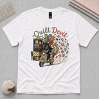A white short-sleeve graphic t-shirt laid flat on a gray textured background, featuring a colorful illustration of a Tasmanian Devil riding a green scooter with a burst of quilt fabric and the text 'Quilt Devil'.