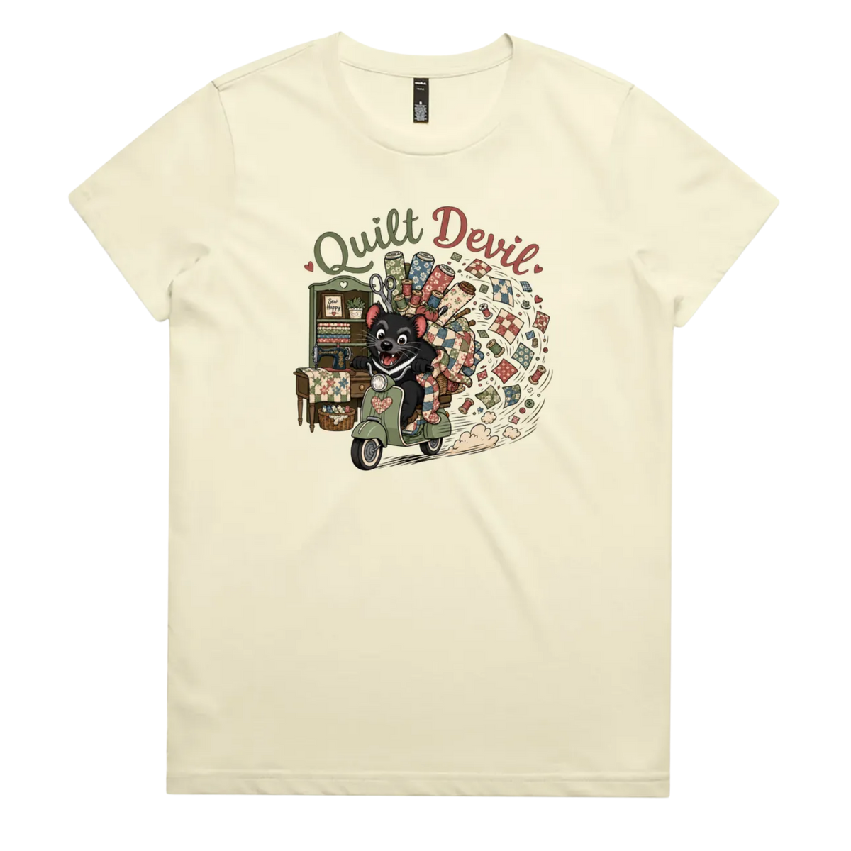 A pale butter yellow short-sleeve crewneck t-shirt featuring a colorful graphic print of a Tasmanian Devil riding a green scooter, surrounded by quilts, fabric and quilting tools with the text 'Quilt Devil'.