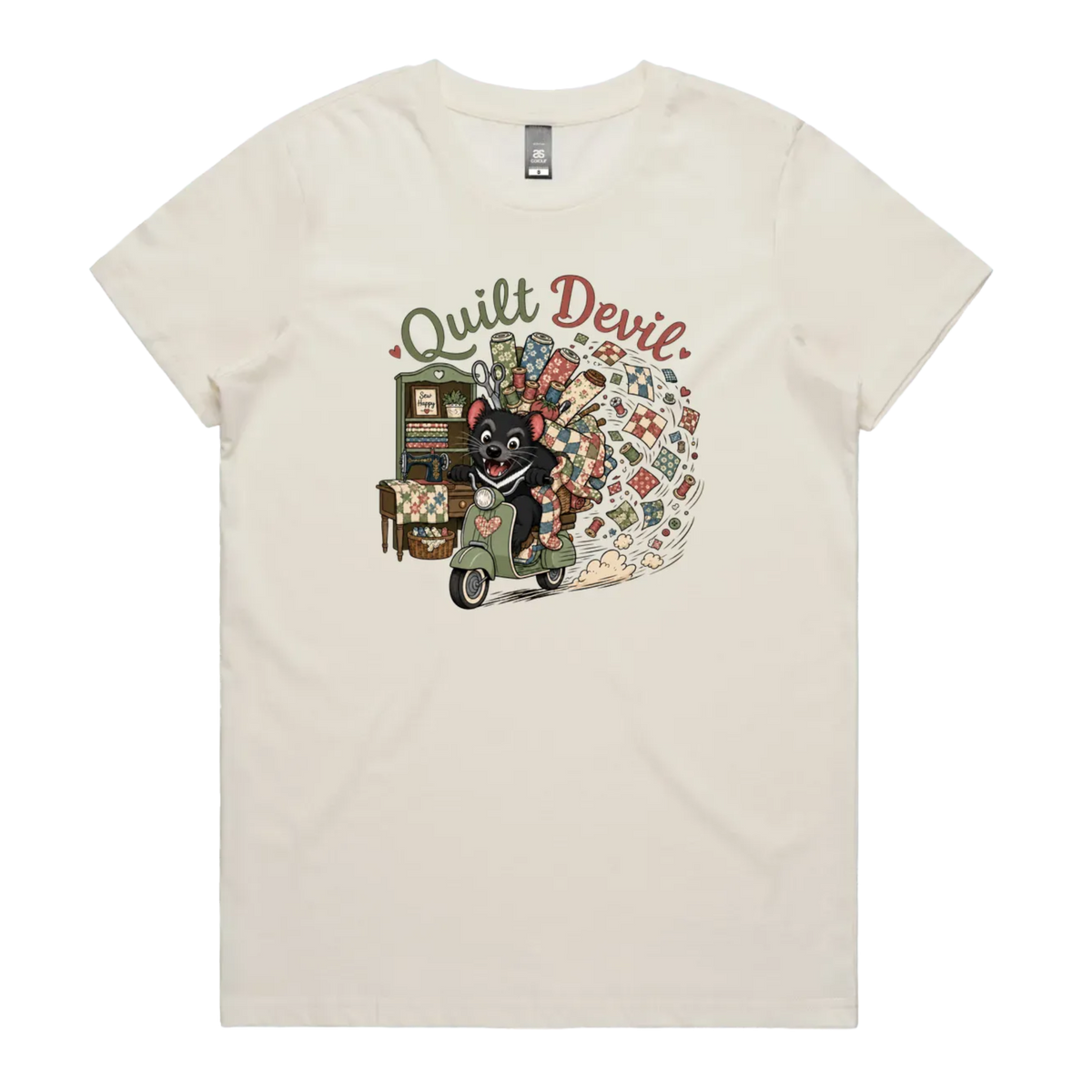 A light cream short-sleeve crewneck t-shirt featuring a colorful graphic print of a Tasmanian Devil riding a green scooter, surrounded by quilts, fabric and quilting tools with the text 'Quilt Devil'.