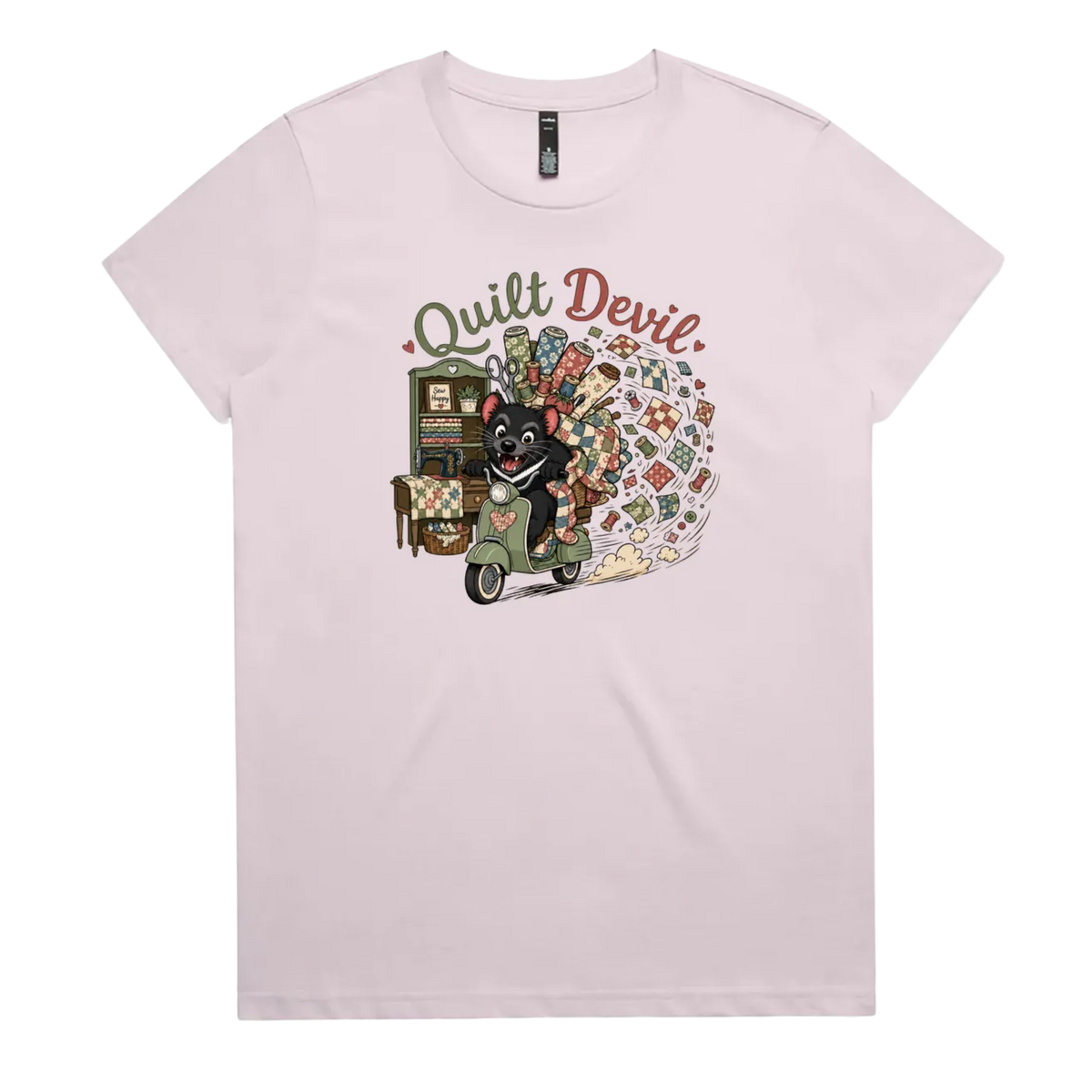 A light mauve short-sleeve crewneck t-shirt featuring a colorful graphic print of a Tasmanian Devil riding a green scooter, surrounded by quilts, fabric and quilting tools with the text 'Quilt Devil'.