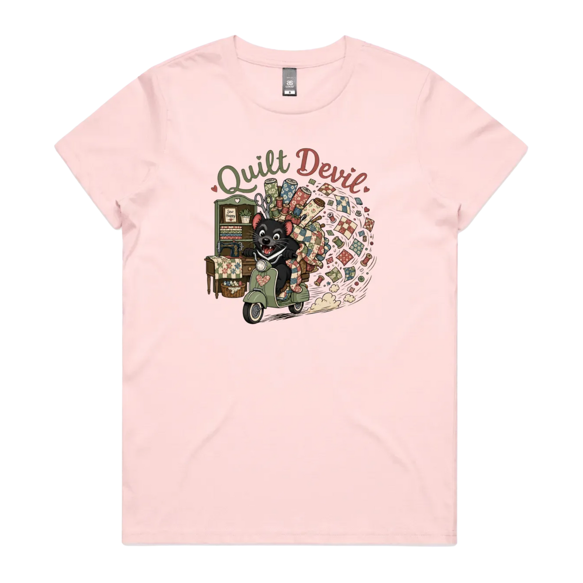 A light pink short-sleeve crewneck t-shirt featuring a colorful graphic print of a Tasmanian Devil riding a green scooter, surrounded by quilts, fabric and quilting tools with the text 'Quilt Devil'.