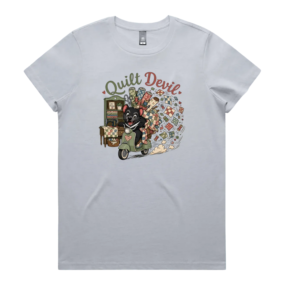 A light powder blue short-sleeve crewneck t-shirt featuring a colorful graphic print of a Tasmanian Devil riding a green scooter, surrounded by quilts, fabric and quilting tools with the text 'Quilt Devil'.