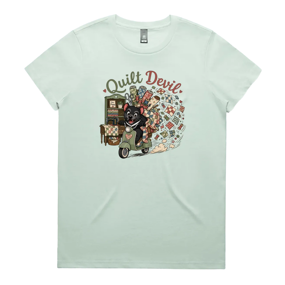 A light seafoam green short-sleeve crewneck t-shirt featuring a colorful graphic print of a Tasmanian Devil riding a green scooter, surrounded by quilts, fabric and quilting tools with the text 'Quilt Devil'.