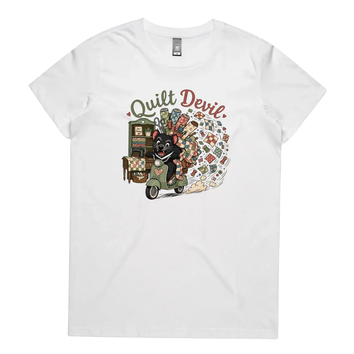 A white short-sleeve crewneck t-shirt featuring a colorful graphic print of a Tasmanian Devil riding a green scooter, surrounded by quilts, fabric and quilting tools with the text 'Quilt Devil'.