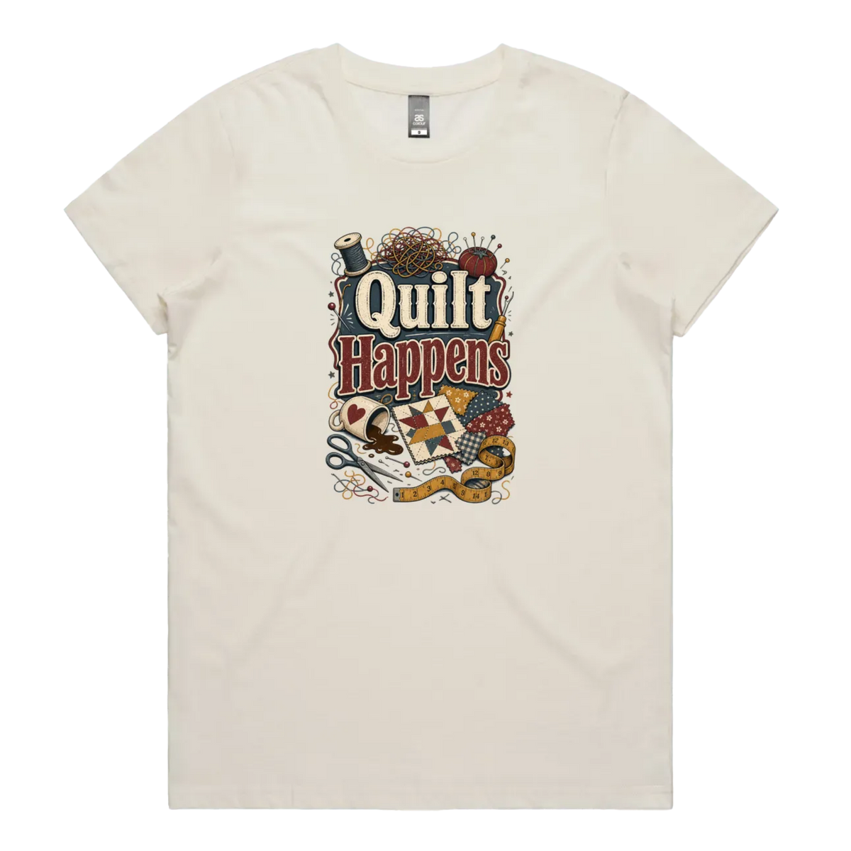 A pale cream short-sleeve crewneck t-shirt featuring a colorful graphic print on the front with the text 'Quilt Happens' surrounded by quilting tools and fabrics.