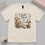 An light cream short-sleeve t-shirt with a vintage-style armchair, a small table with coffee cup and the text 'Quilt More Sip Slowly' in a floral arrangement. The shirt is displayed flat on a pale grey cutting mat with quilting props, including thread spools and scissors.