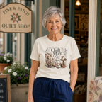 An older woman with short grey hair standing outside a quilt shop wearing a light cream graphic t-shirt featuring the words 'Quilt More, Sip Slowly'.