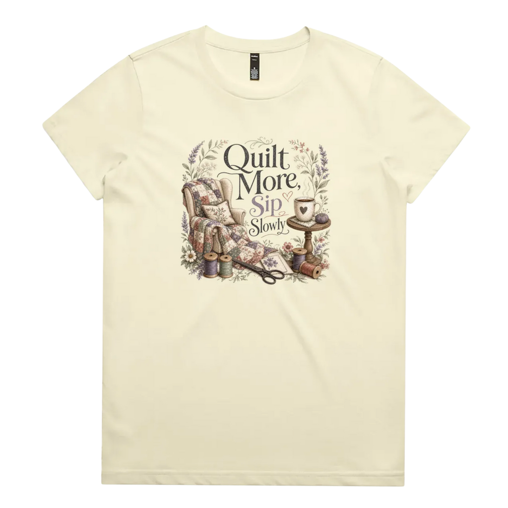 A pale butter yellow short-sleeve crewneck t-shirt featuring a vintage-style graphic on the front with the text 'Quilt More, Sip Slowly' surrounding an illustration of a quilted armchair, floral details, and sewing tools.