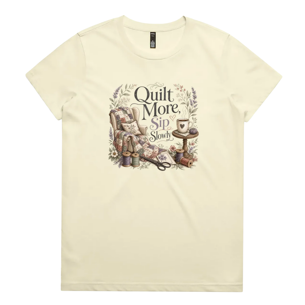 A pale butter yellow short-sleeve crewneck t-shirt featuring a vintage-style graphic on the front with the text 'Quilt More, Sip Slowly' surrounding an illustration of a quilted armchair, floral details, and sewing tools.