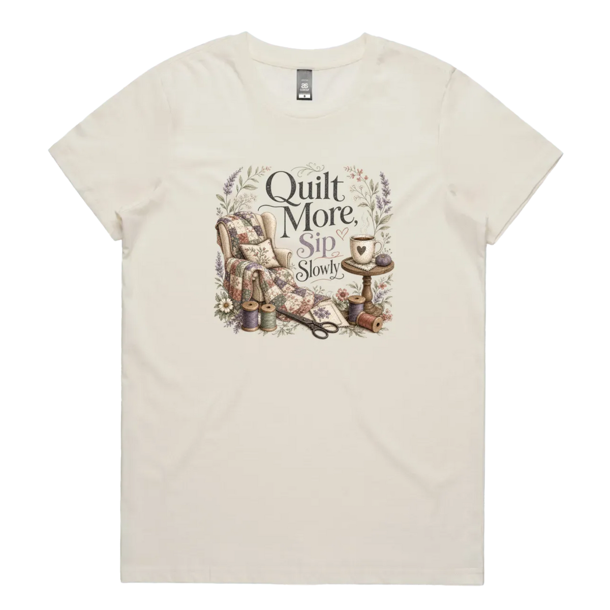 A pale cream short-sleeve crewneck t-shirt featuring a vintage-style graphic on the front with the text 'Quilt More, Sip Slowly' surrounding an illustration of a quilted armchair, floral details, and sewing tools.