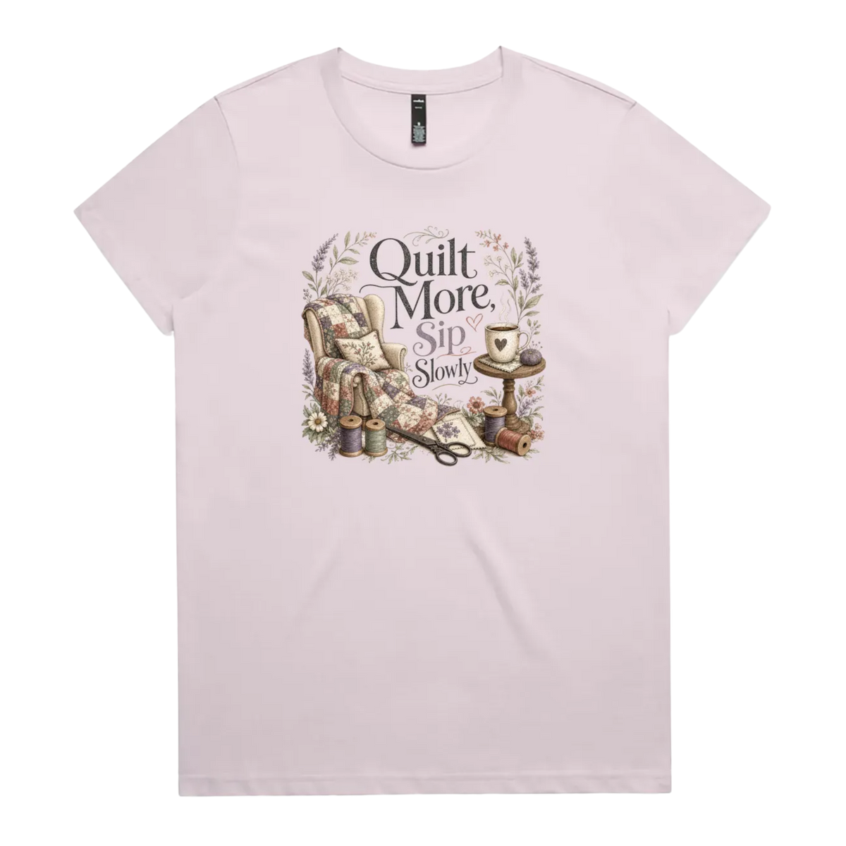 A pale mauve short-sleeve crewneck t-shirt featuring a vintage-style graphic on the front with the text 'Quilt More, Sip Slowly' surrounding an illustration of a quilted armchair, floral details, and sewing tools.
