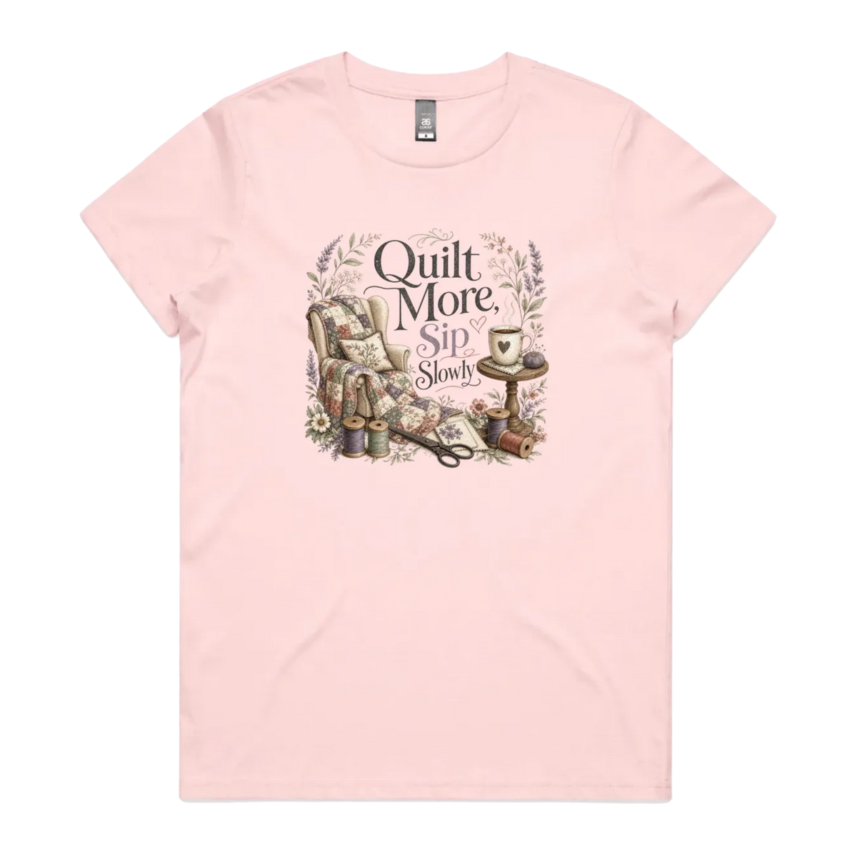A pale pink short-sleeve crewneck t-shirt featuring a vintage-style graphic on the front with the text 'Quilt More, Sip Slowly' surrounding an illustration of a quilted armchair, floral details, and sewing tools.