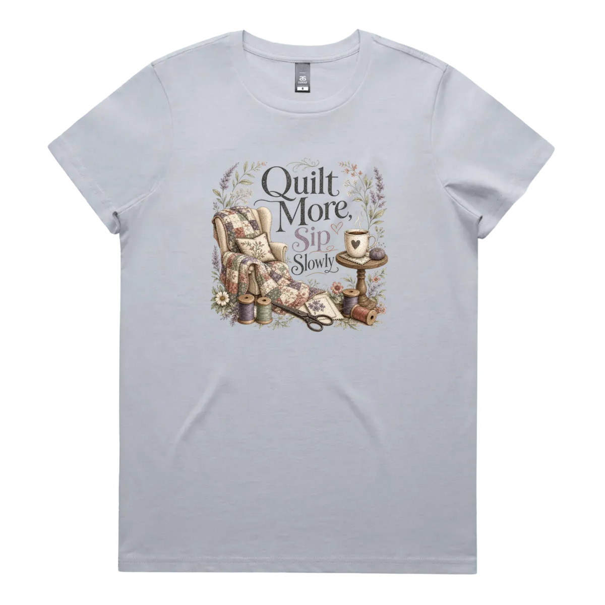 A pale powder blue short-sleeve crewneck t-shirt featuring a vintage-style graphic on the front with the text 'Quilt More, Sip Slowly' surrounding an illustration of a quilted armchair, floral details, and sewing tools.
