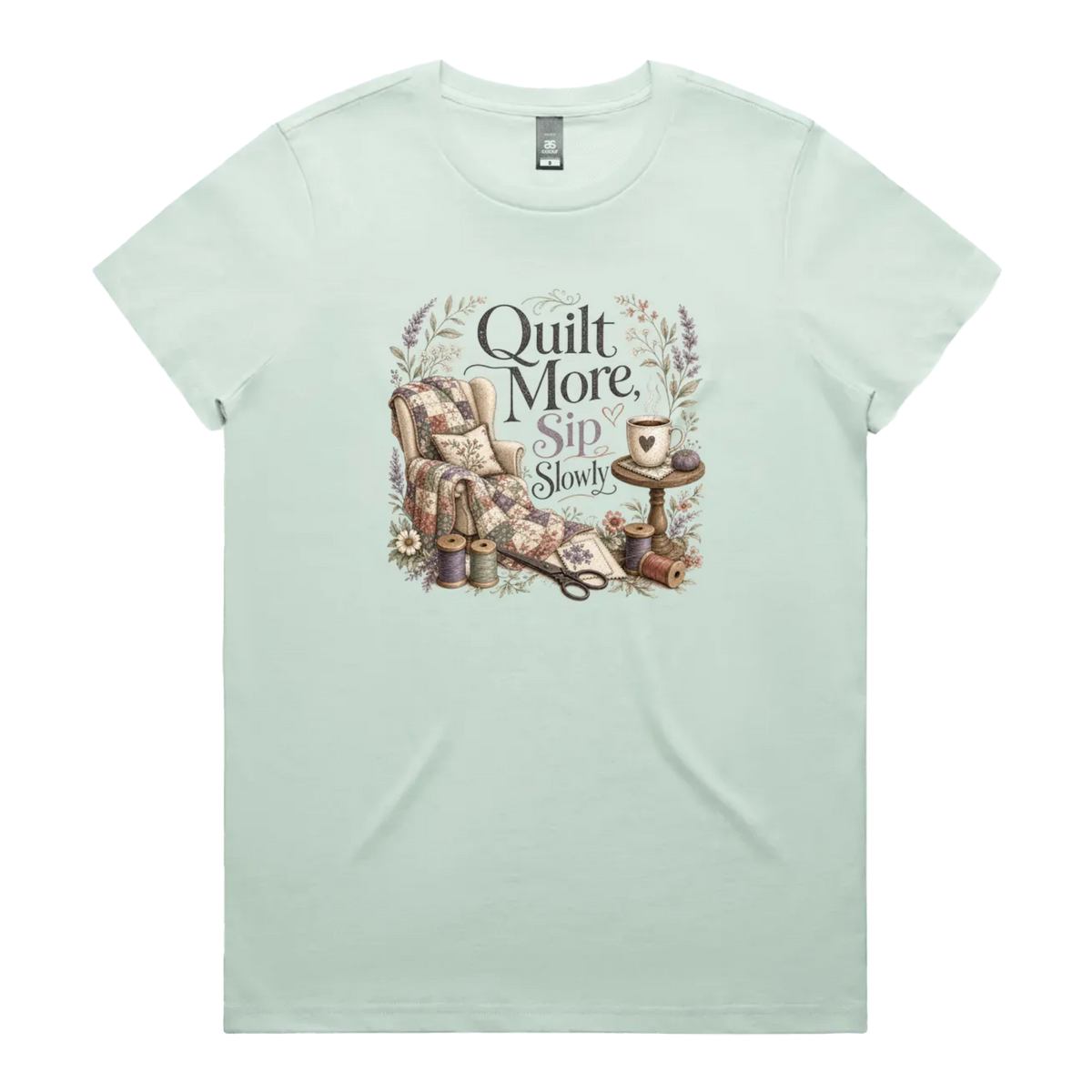 A pale seafoam green short-sleeve crewneck t-shirt featuring a vintage-style graphic on the front with the text 'Quilt More, Sip Slowly' surrounding an illustration of a quilted armchair, floral details, and sewing tools.