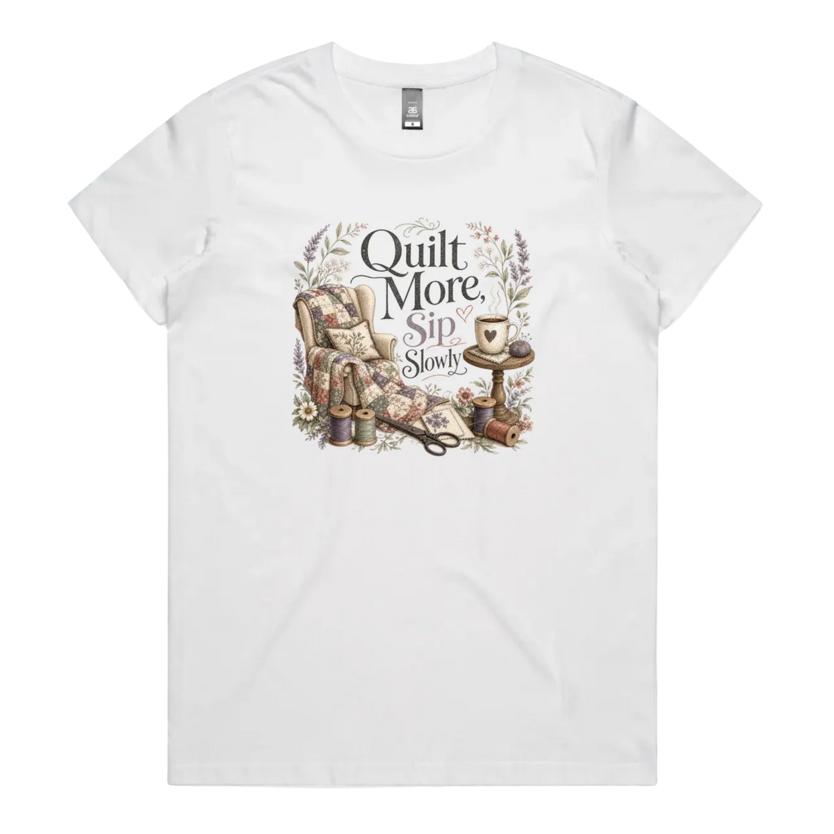 A white short-sleeve crewneck t-shirt featuring a vintage-style graphic on the front with the text 'Quilt More, Sip Slowly' surrounding an illustration of a quilted armchair, floral details, and sewing tools.