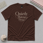 A brown short-sleeve crewneck t-shirt with the text 'Quietly Making Things' in a cream script font on the chest. The shirt is laid flat on a gray curring mat with a stack of folded fabric, a quilting ruler and a sheep pincushion in the corners.