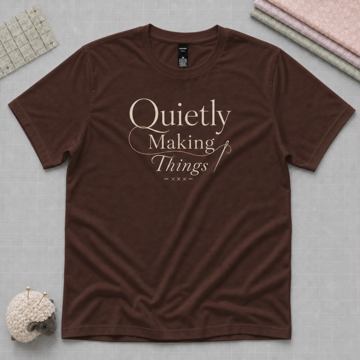 A brown short-sleeve crewneck t-shirt with the text 'Quietly Making Things' in a cream script font on the chest. The shirt is laid flat on a gray curring mat with a stack of folded fabric, a quilting ruler and a sheep pincushion in the corners.