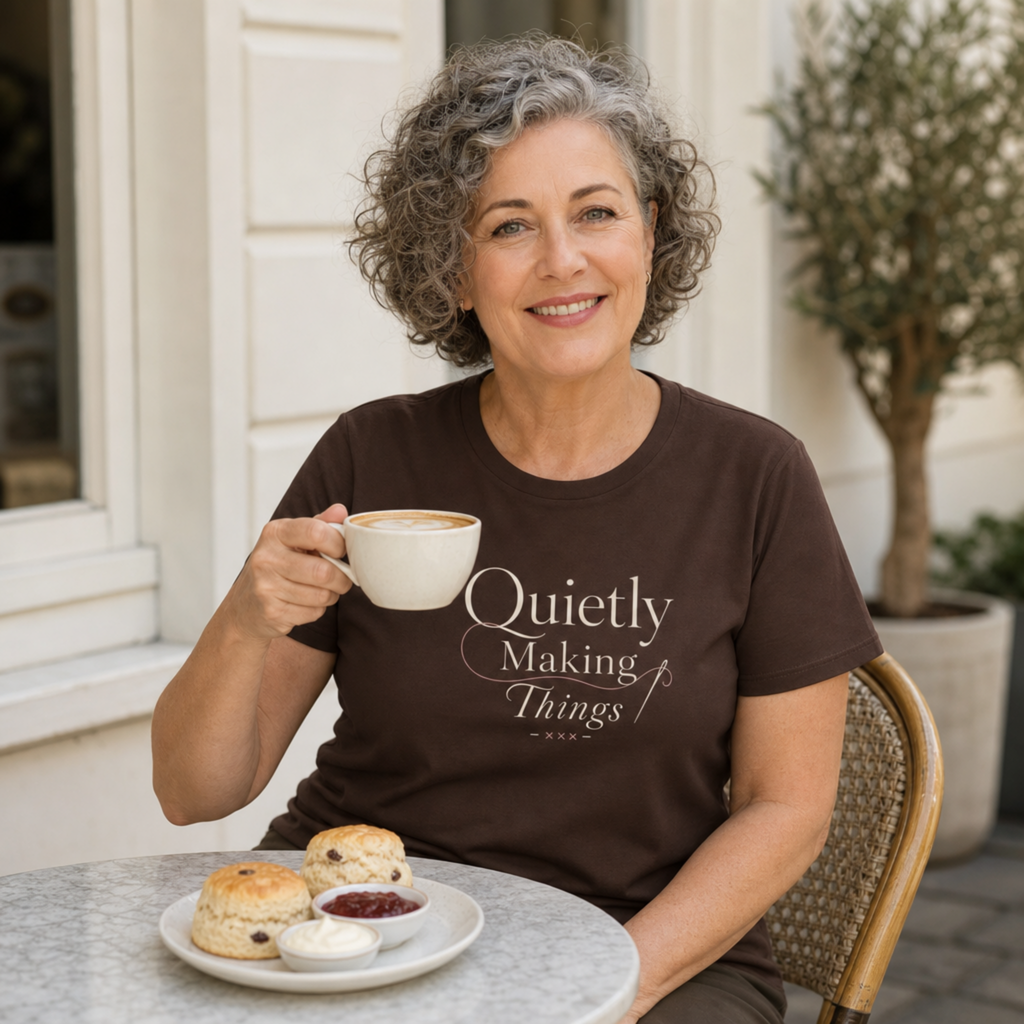 A smiling woman with gray curly hair sits at an outdoor cafe table wearing a dark brown t-shirt with the text 'Quietly Making Things' in cream script. She holds a white coffee mug, with a plate of scones and cream on the table in front of her.