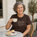 A smiling woman with gray curly hair sits at an outdoor cafe table wearing a dark brown t-shirt with the text 'Quietly Making Things' in cream script. She holds a white coffee mug, with a plate of scones and cream on the table in front of her.