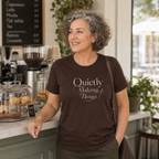 A woman with curly hair standing at a cafe counter wearing a dark brown t-shirt with the words 'Quietly Making Things' in cream script.