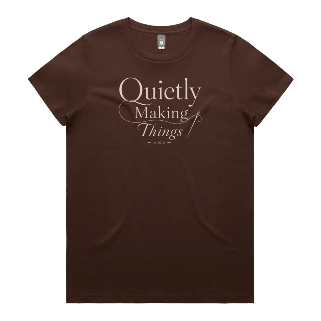A dark brown short-sleeve crewneck t-shirt featuring a cream-colored graphic print on the front that reads 'Quietly Making Things' in an elegant serif and script font with needle and thread accents.