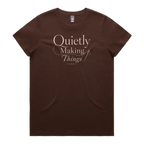 A dark brown short-sleeve crewneck t-shirt featuring a cream-colored graphic print on the front that reads 'Quietly Making Things' in an elegant serif and script font with needle and thread accents.