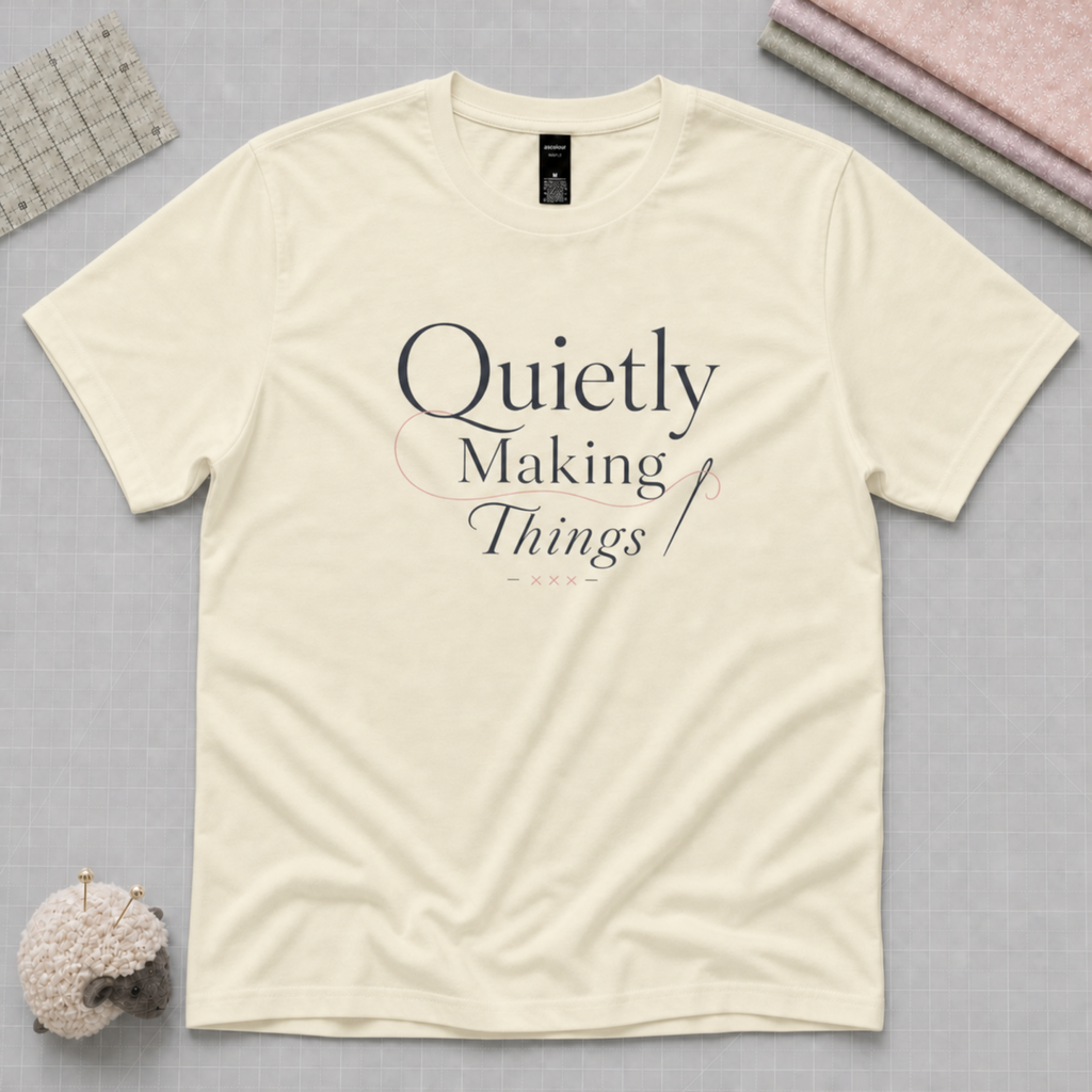 A pale butter yellow short-sleeve t-shirt with the text 'Quietly Making Things' printed on the front, styled in a flat-lay with sewing and crafting props on a gray background.