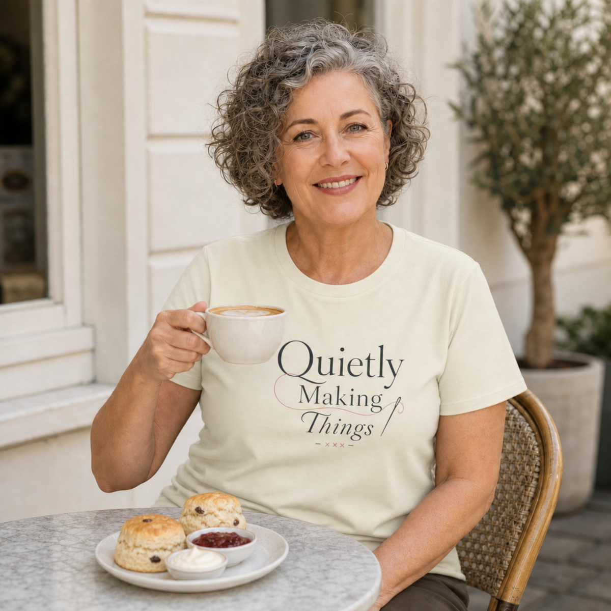 A smiling woman with gray curly hair sits at an outdoor cafe table holding a mug, wearing a cream-colored t-shirt with the text 'Quietly Making Things' printed on the front. In front of her is a plate with scones and cream.