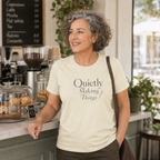 A smiling woman with curly gray hair stands in a cafe wearing a cream-colored t-shirt with the printed text 'Quietly Making Things'.