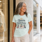 A woman with long grey hair standing in a mall wearing a pale mint green t-shirt with the graphic 'Queen of the Quilt Frame'.