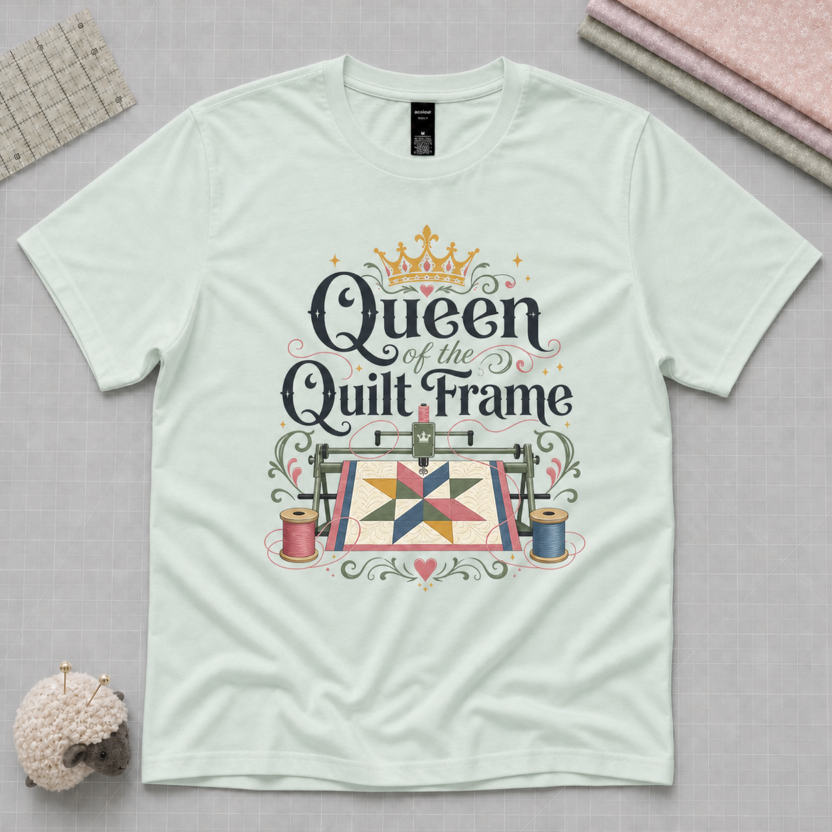 A pale mint green short-sleeve t-shirt laying flat on a pale background. The graphic on the t-shirt reads 'Queen of the Quilt Frame' above an illustration of a longarm machine, quilt and thread spools.