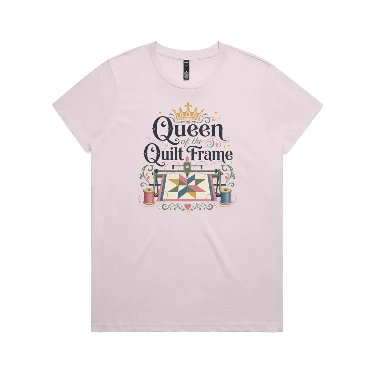 A pale mauve short-sleeve t-shirt with a graphic print on the front that reads 'Queen of the Quilt Frame' in black script, decorated with a gold crown, a quilt design, and spools of thread.