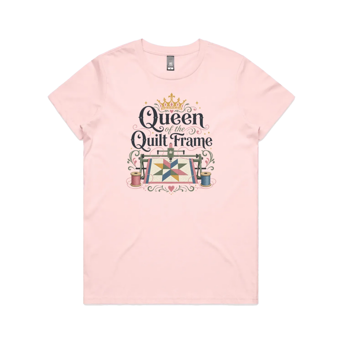 A pale pink short-sleeve t-shirt with a graphic print on the front that reads 'Queen of the Quilt Frame' in black script, decorated with a gold crown, a quilt design, and spools of thread.