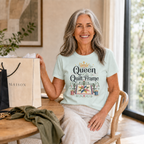 A smiling grey haired woman sitting at a table wearing the graphic t-shirt 'Queen of the Quilt Frame'. There are shopping bags on the table.