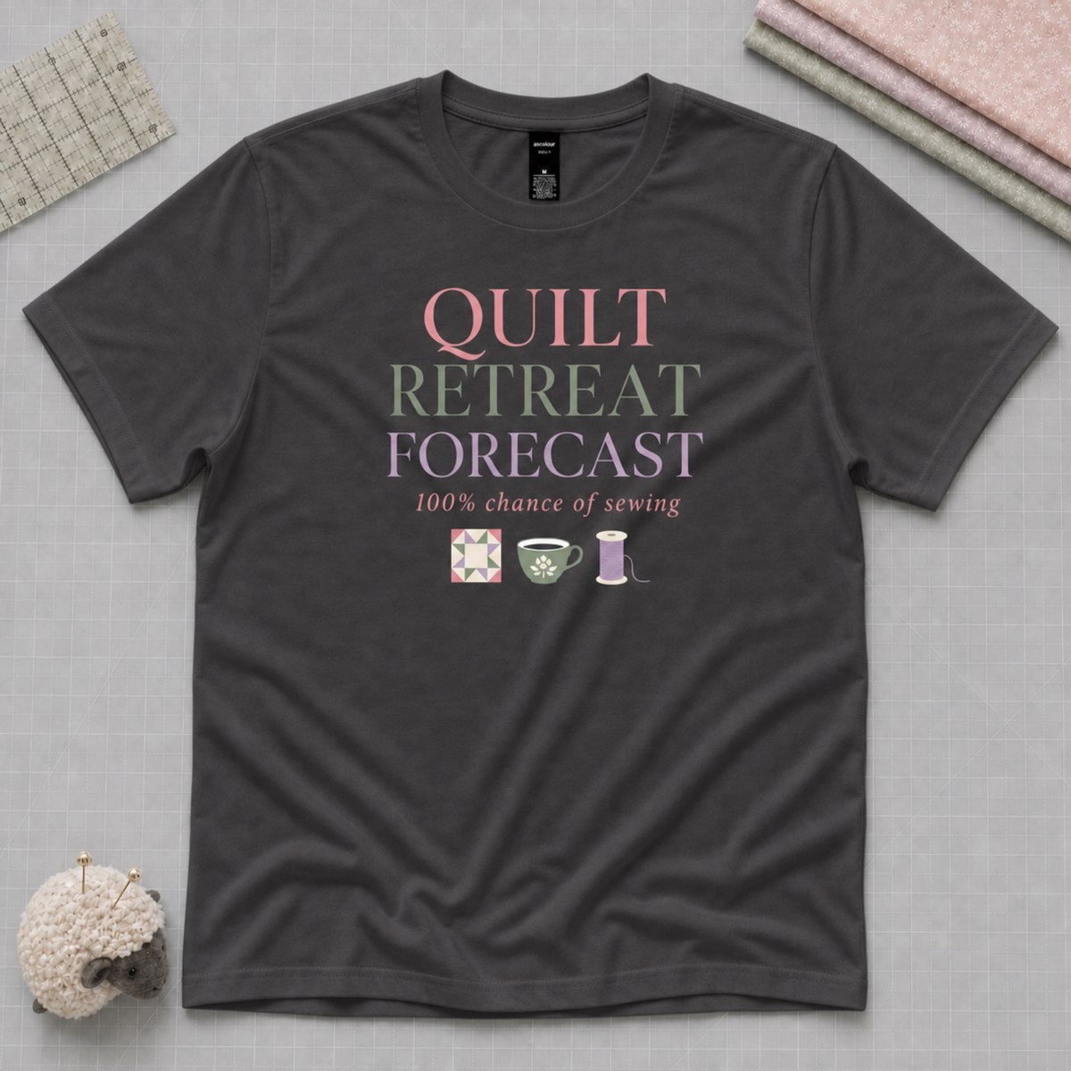 A charcoal grey short-sleeve t-shirt with the words 'Quilt Retreat Forecast, 100% chance of sewing' in pink, green and mauve with small quilt motifs. The shirt lays flat on a pale background with quilt themed accessories in the corners.