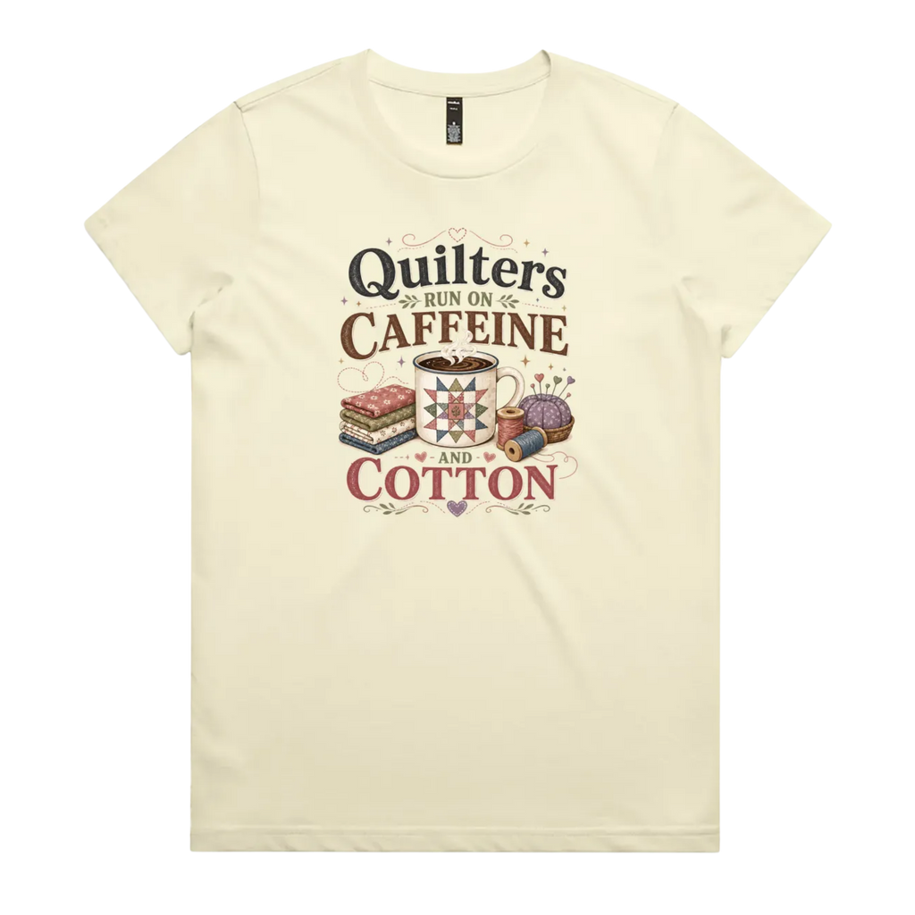 A light butter yellow short-sleeve crewneck t-shirt featuring a retro-style graphic print on the front. The graphic reads 'Quilters Run on Caffeine and Cotton' in stylized fonts, accompanied by illustrations of a fabric stack, a mug of coffee, a pincushion, and a spool of thread.