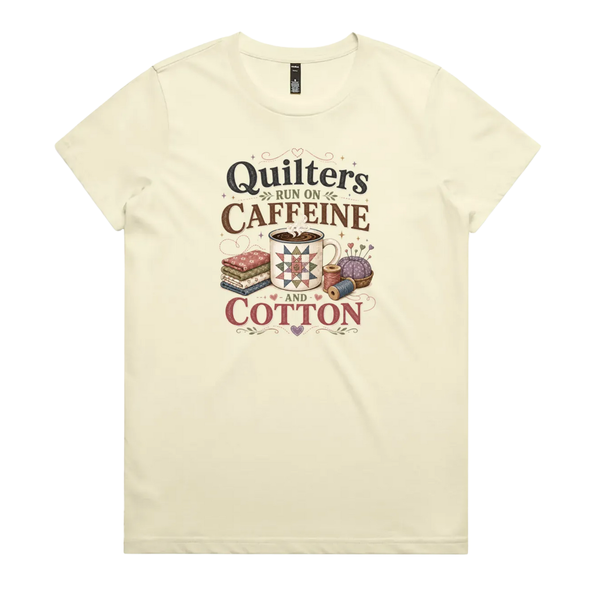 A light butter yellow short-sleeve crewneck t-shirt featuring a retro-style graphic print on the front. The graphic reads 'Quilters Run on Caffeine and Cotton' in stylized fonts, accompanied by illustrations of a fabric stack, a mug of coffee, a pincushion, and a spool of thread.