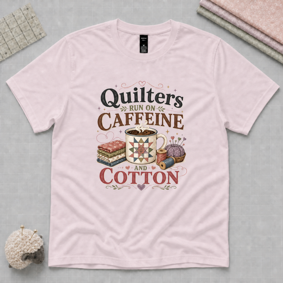 A light mauve short-sleeve t-shirt with a vintage-style graphic that reads 'Quilters Run on Caffeine and Cotton' with illustrations of a coffee mug, fabric stack, and sewing notions. The shirt is displayed flat on a light grey cutting mat with folded fabrics, a quilting ruler and sheep pincushion in the corners.