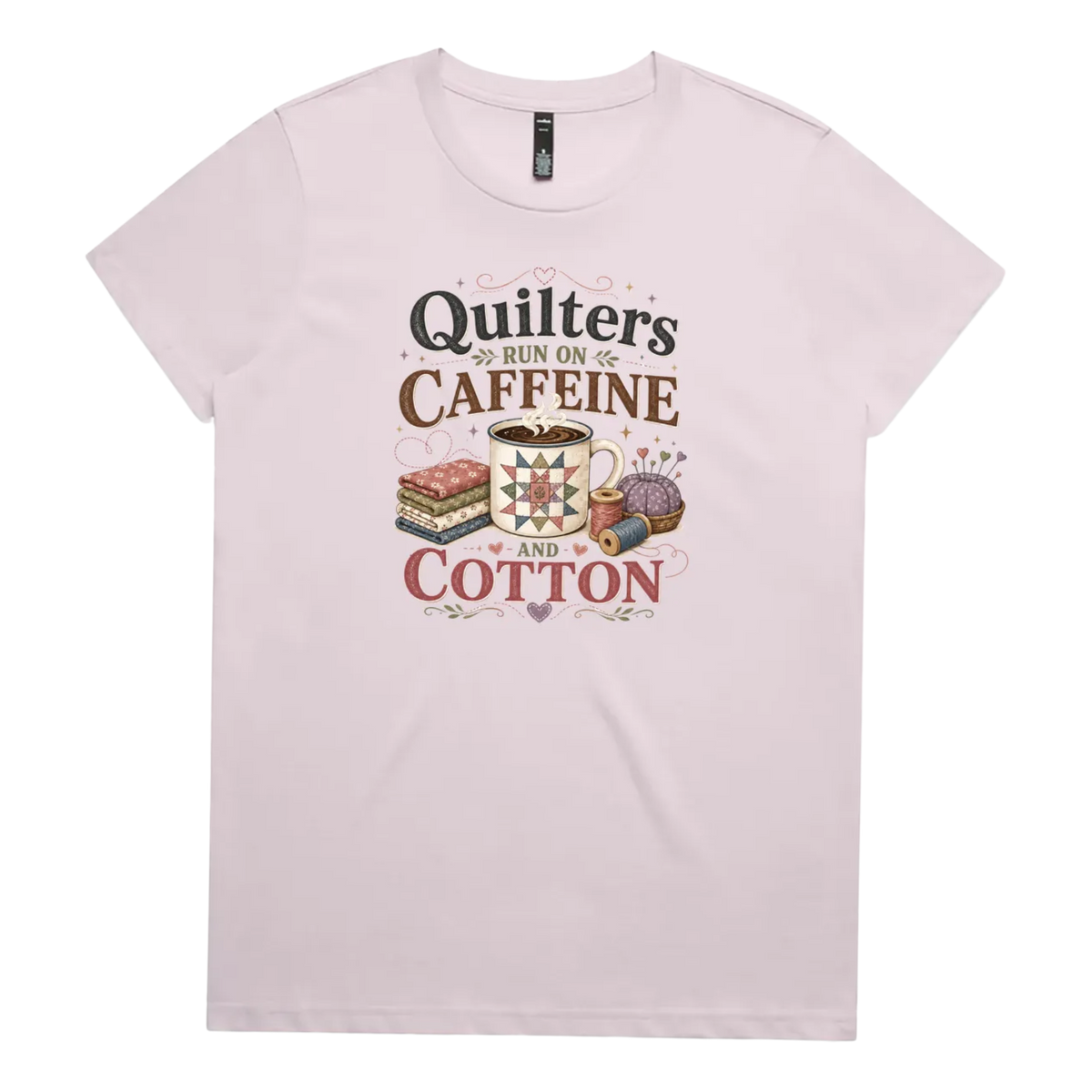 A light mauve short-sleeve crewneck t-shirt featuring a retro-style graphic print on the front. The graphic reads 'Quilters Run on Caffeine and Cotton' in stylized fonts, accompanied by illustrations of a fabric stack, a mug of coffee, a pincushion, and a spool of thread.
