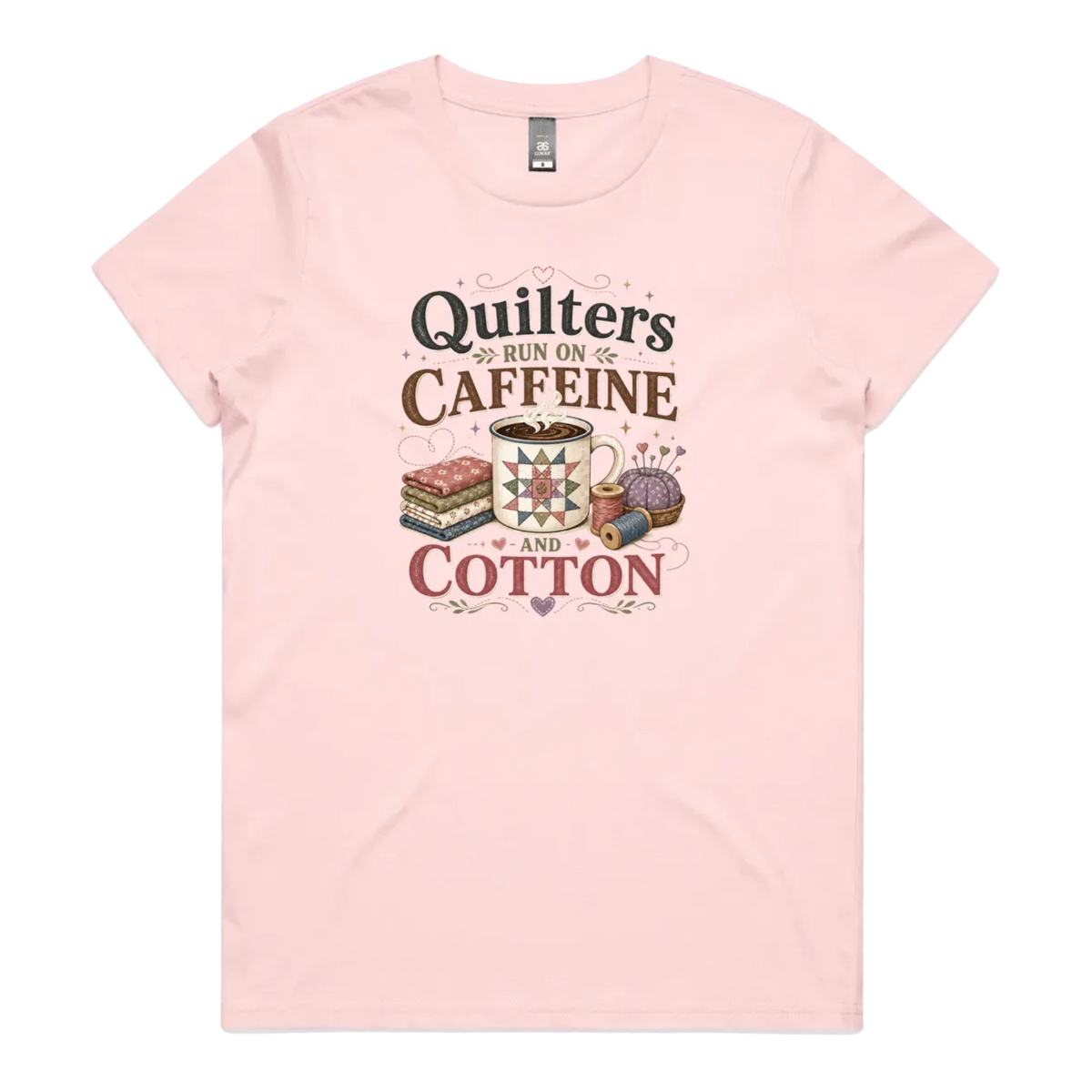 A light pink short-sleeve crewneck t-shirt featuring a retro-style graphic print on the front. The graphic reads 'Quilters Run on Caffeine and Cotton' in stylized fonts, accompanied by illustrations of a fabric stack, a mug of coffee, a pincushion, and a spool of thread.