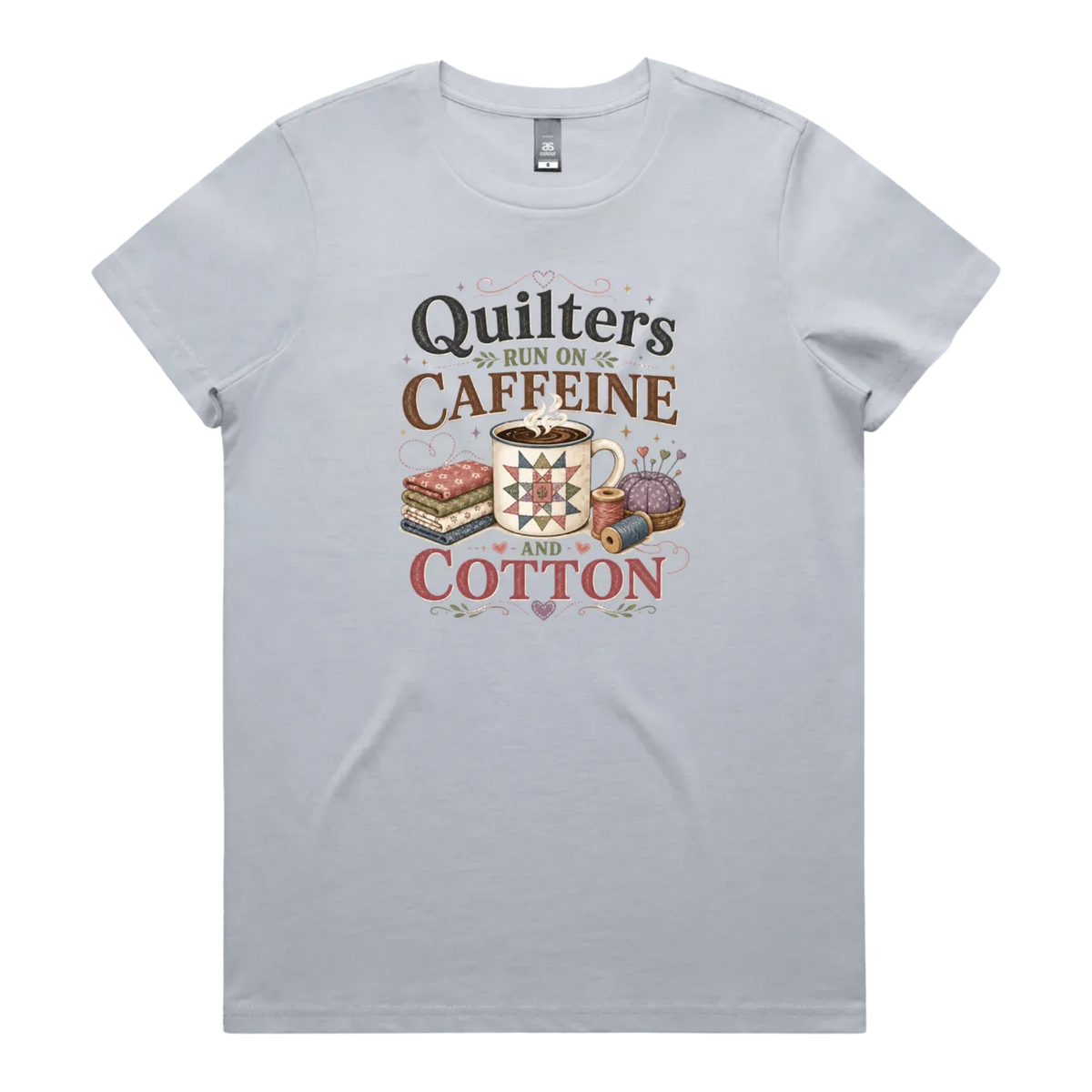 A light powder blue short-sleeve crewneck t-shirt featuring a retro-style graphic print on the front. The graphic reads 'Quilters Run on Caffeine and Cotton' in stylized fonts, accompanied by illustrations of a fabric stack, a mug of coffee, a pincushion, and a spool of thread.