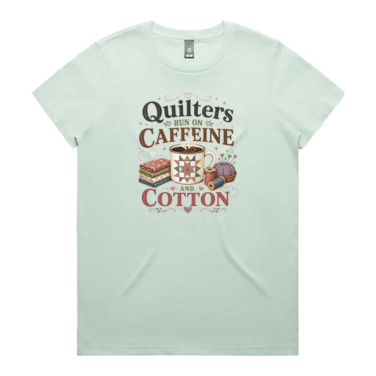 A light seafoam green short-sleeve crewneck t-shirt featuring a retro-style graphic print on the front. The graphic reads 'Quilters Run on Caffeine and Cotton' in stylized fonts, accompanied by illustrations of a fabric stack, a mug of coffee, a pincushion, and a spool of thread.